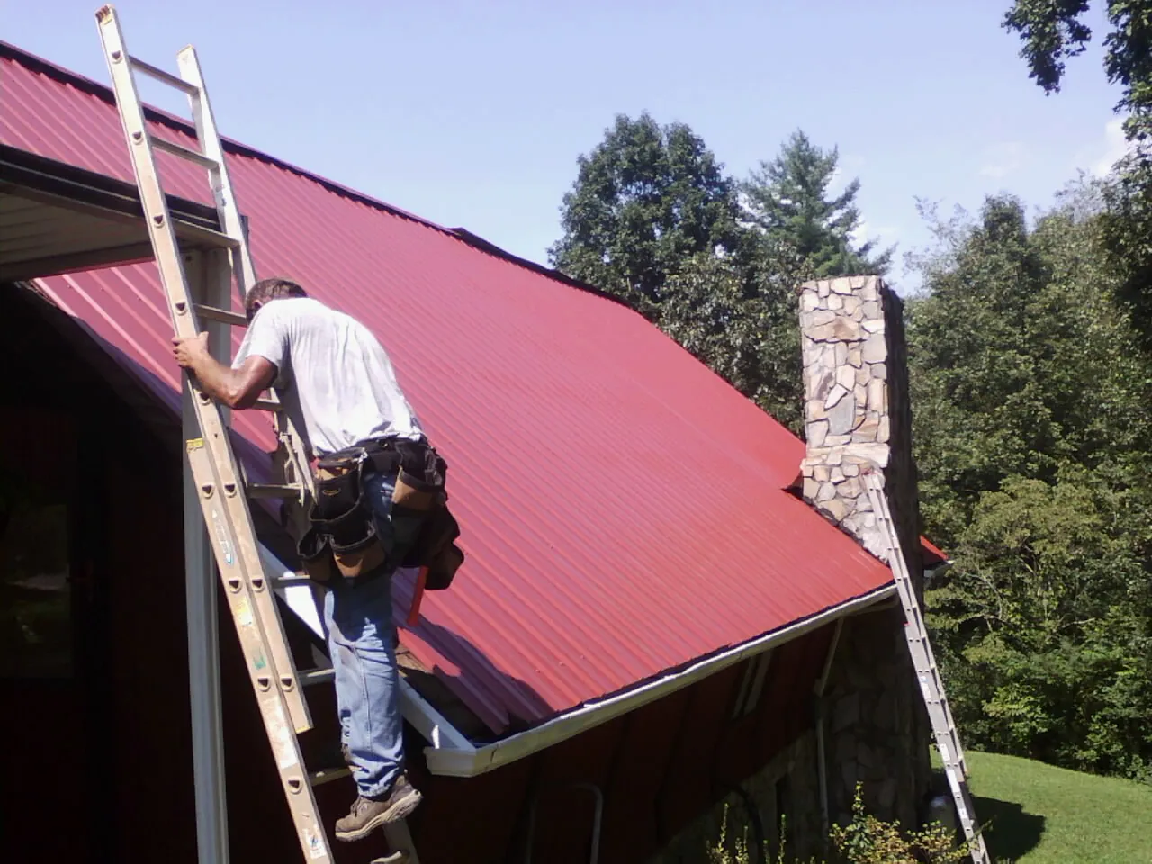 Roofer on a ladder beside a red metal roof and stone chimney — Soffit & Fascia Repair by Roofing Brothers in Palo Cedro